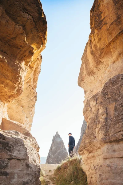 El hombre de ropa oscura y gorra de béisbol se encuentra entre hermosas rocas y admira el paisaje en Capadocia en Turquía —  Fotos de Stock