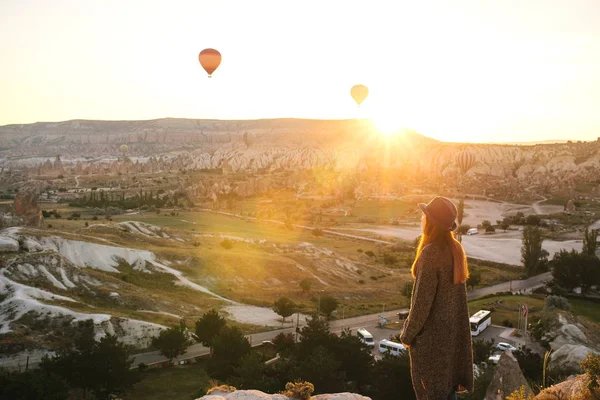 A tourist girl in a hat admires hot air balloons flying in the sky over Cappadocia in Turkey. Impressive sight. — Stock Photo, Image