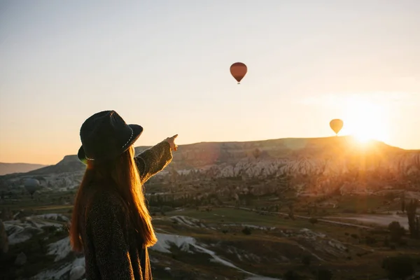 A tourist girl in a hat admires hot air balloons flying in the sky over Cappadocia in Turkey. Impressive sight. — Stock Photo, Image