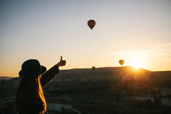 A tourist girl in a hat admires hot air balloons flying in the sky over Cappadocia in Turkey. Impressive sight. — Stock Photo, Image