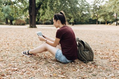Bir sırt çantası ile genç bir kız sonbahar parkta oturur ve tablet kullanır.