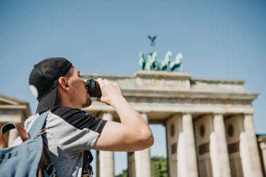 Bir kişi tek kullanımlık bir fincan kahve ya da Berlin Brandenburg Kapısı'nın arka planda başka bir içecek ile tutar.