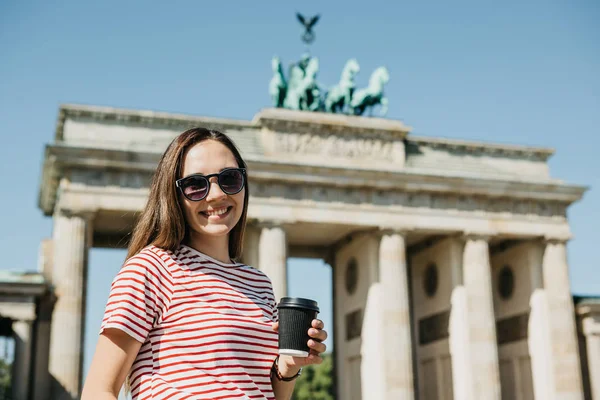 Una persona tiene una tazza usa e getta con caffè o un altro drink sullo sfondo della Porta di Brandeburgo a Berlino . — Foto Stock