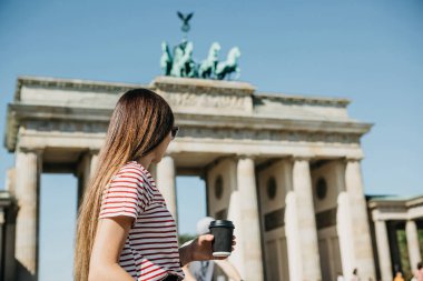 Bir kişi tek kullanımlık bir fincan kahve ya da Berlin Brandenburg Kapısı'nın arka planda başka bir içecek ile tutar.