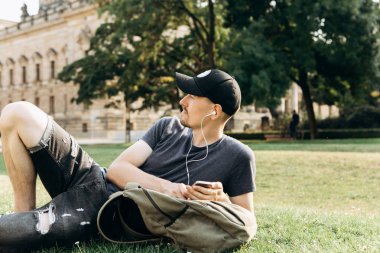 Bir sırt çantası ya da bir öğrenci olan genç bir adam çimenlerin üzerinde yatıyor ve müzik veya bir podcast için dinler ve sahiptir.