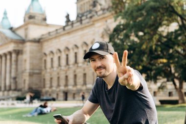 Genç adam turist veya öğrenci Almanya'da Leipzig, sokaklarında çimenlerin üzerinde oturan ve el hareketi barış demek gösterir