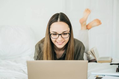 Güzel, gülümseyen, pozitif kız evde yatarken dizüstü bilgisayar kullanıyor..