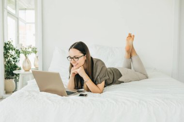 Pozitif kız öğrenci yatakta yatar ve dizüstü bilgisayar kullanır. Ekrandan kitap okuyor ya da video veya eğitim kursu izliyor..