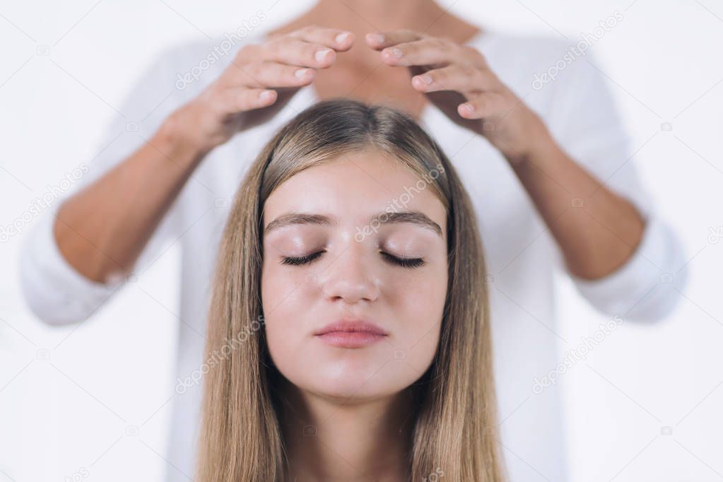Horizontal image of female Reiki therapist standing and holding hands above head of the beautiful teenage patient. Reiki Practitioner transfering energy and healing crown chakra. Peaceful girl sitting with her eyes closed