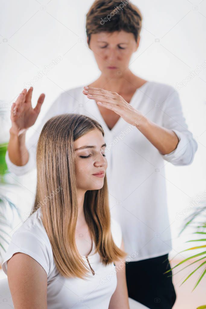 Horizontal image of female Reiki therapist standing and holding hands above head of the beautiful teenage patient. Reiki Practitioner transfering energy and healing crown chakra. Peaceful girl sitting with her eyes closed