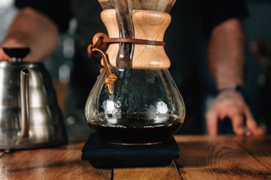 Filtre kahve makinesi, termometre ve ahşap masa üzerinde dijital ölçek su ısıtıcısı, yakın çekim. Barista dövmeli silah karanlık üniforması ile.    