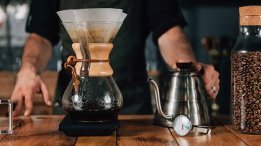 Filtre kahve makinesi, termometre ve ahşap masa üzerinde dijital ölçek su ısıtıcısı, yakın çekim. Barista dövmeli silah karanlık üniforması ile.    