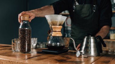 Filtre kahve makinesi, termometre ve ahşap masa üzerinde dijital ölçek su ısıtıcısı, yakın çekim. Barista dövmeli silah karanlık üniforması ile.    