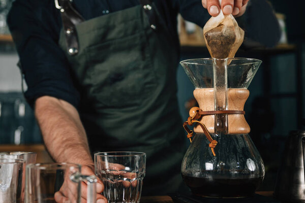 Портрет молодого бариста, улыбающегося. Бариста с татуированными руками в тёмной форме. Инструменты и оборудование для приготовления фильтра кофе на столе
.