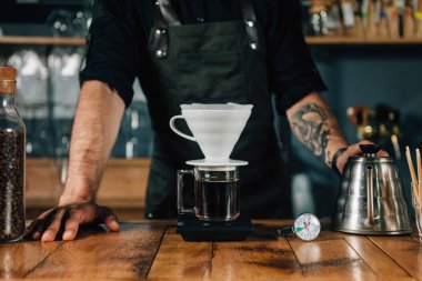 Filtre kahve makinesi, termometre ve ahşap masa üzerinde dijital ölçek su ısıtıcısı, yakın çekim. Araçları ve ekipmanları damla Brew Kahve yapmak için. Barista dövmeli silah karanlık üniforması ile.    