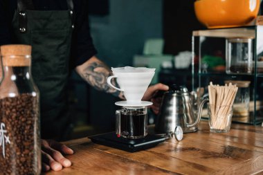Filtre kahve makinesi, termometre ve ahşap masa üzerinde dijital ölçek su ısıtıcısı, yakın çekim. Araçları ve ekipmanları damla Brew Kahve yapmak için. Barista dövmeli silah karanlık üniforması ile.    