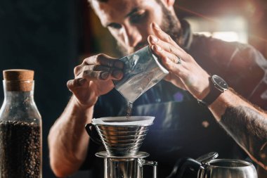 Close up of a young male barista pouring coffee into Kalita Wave Dripper to brew coffee and measure it on digital scale in coffee shop. Barista wearing dark uniform. Tools and equipment for making Drip Brew coffee on table. clipart