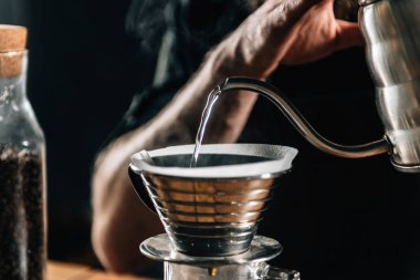 Close up of a young male barista pouring boiling water into Kalita Wave Dripper to brew coffee and measure it on digital scale in coffee shop. Barista wearing dark uniform. Tools and equipment for making Drip Brew coffee on table. clipart