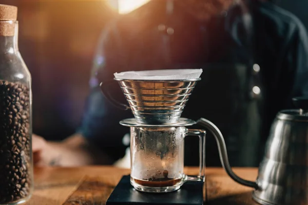 Close up Kalita Wave Dripper on black digital scale, wooden table. Tools and equipment for making Drip Brew coffee. Barista with tattooed arms wearing dark uniform.