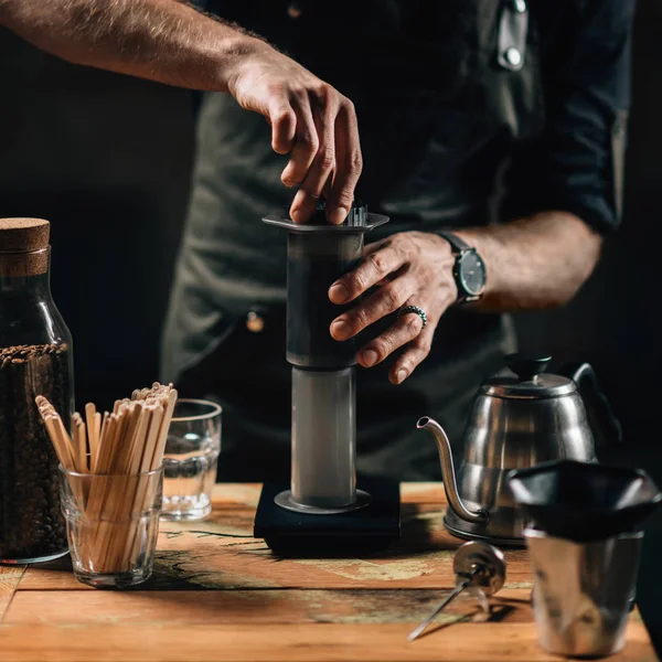 Close up square image of barista making air press coffee. Barista with tattooed arms wearing dark uniform.