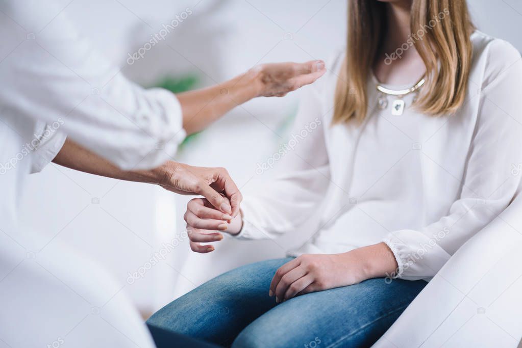 Female Theta healing therapist performing alternative therapy treatment with young woman patient. Therapist doing applied and muscle testing by holding hands. Wearing white clothes.