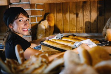 Taze pişmiş baget ile sıcak fırın tepsisi tutarak siyah üniformalı gülümseyen kadın fırın işçisi giyinmiş.