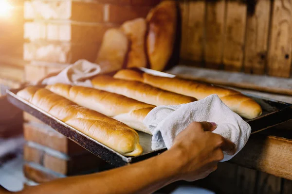 Hands of female baker holding hot baking tray with freshly baked baguettes.