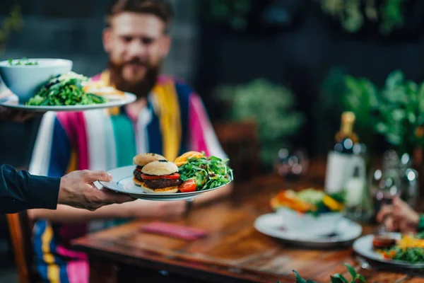 Horizontal image of male hand waiter serving plate of vegetarian burger to a young man in restaurant.