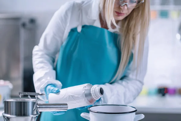 Female candle maker pouring paraffin into the pot with melted wax. Wearing protective work wear. Candle making process