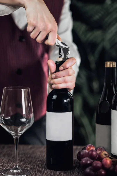 女侍酒师打开酒瓶用软木塞 桌子上的新鲜葡萄和空葡萄酒杯 — 图库照片