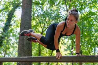 An athletic woman showcases her agility, leaping over an outdoor wooden barrier in a park.