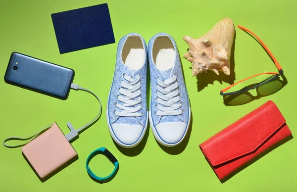 Women\'s trendy travel accessories and technology gadgets on the green surface. Sneakers, passport, purse, glasses, smartphone, power bank, shell, smart bracelet. Top view. Flat lay.