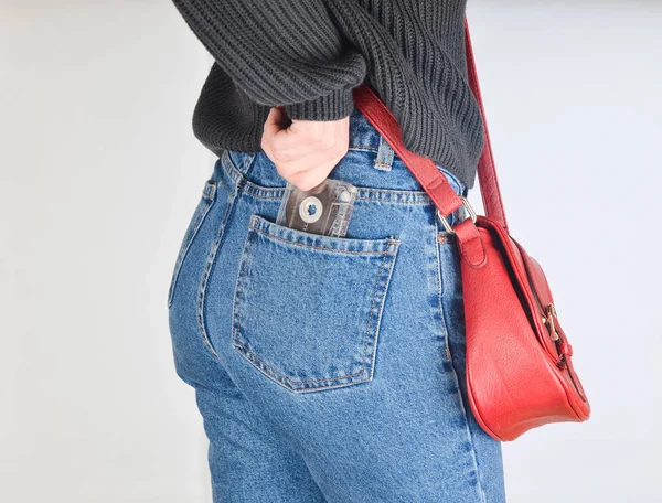 A girl in a mimes fit jeans, a sweater and with a red bag on the edge pulls out a retro audio cassette from her pocket on a white background. Side view.