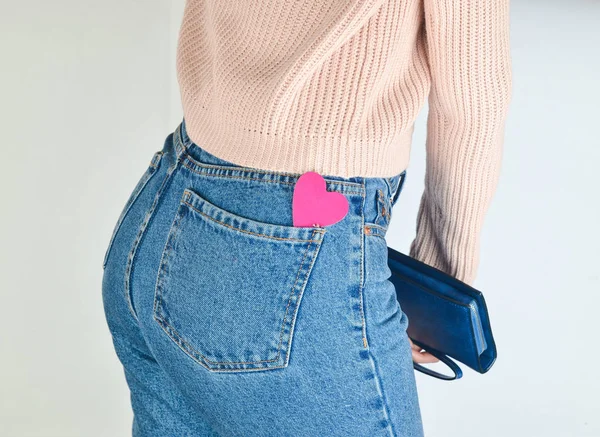 Decorative heart in the back pockets of jeans of a slim girl who holds a purse in her hands on a white background. Side view.