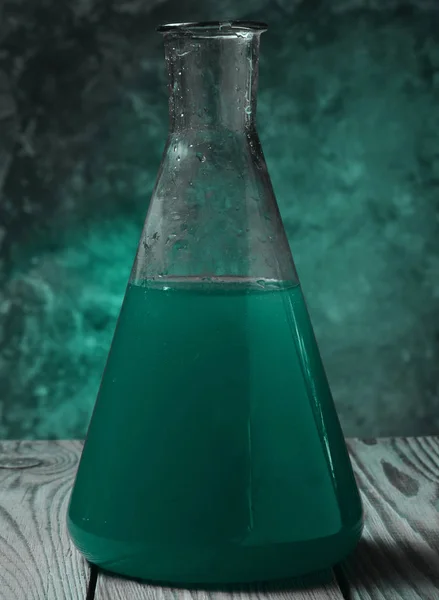 A laboratory flask with a blue liquid on a wooden shelf against a concrete wall.