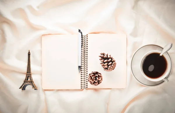 Morning inspiration, cup of tea, notebook with a pen, pine cone, souvenir on a white bed sheet. Breakfast on the bed. The concept of dreams and the desire to travel. Top view.