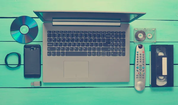 Modern and obsolete devices and gadgets on a turquoise wooden table. Laptop, audio and video cassette, smartphone, remote control, CDs, USB flash drive, smart bracelet.Digital and analog technologies.