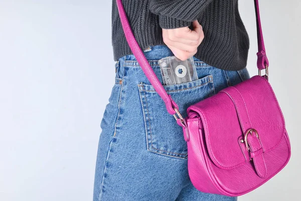 A girl in a mimes fit jeans, a sweater and with a red bag on the edge pulls out a retro audio cassette from her pocket on a white background. Side view.