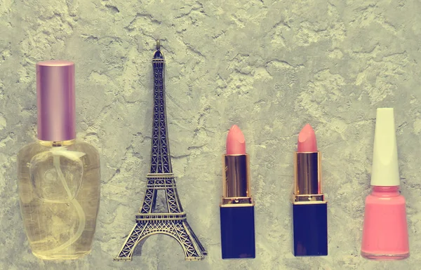 Women\'s cosmetics from Paris. Two pink lipsticks, a perfume bottle, nail polish, a statuette of the Eiffel tower on a gray concrete background. Top view