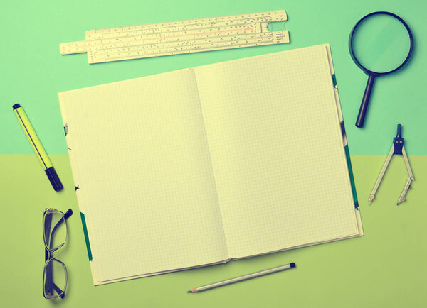 A large notebook for notes and drafting with stationery objects for a student on a green blue pastel background. Ruler, marker, pencil, magnifying glass, compasses, glasses. Top view.