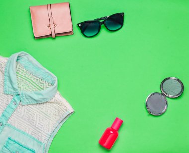 Kadın aksesuarları ve giyim yeşil bir arka plan üzerinde. Kot gömlek, ayna, parfüm şişesi, güneş gözlüğü, çanta. Uzay kopyalayın. Düz yatıyordu. Üstten Görünüm.