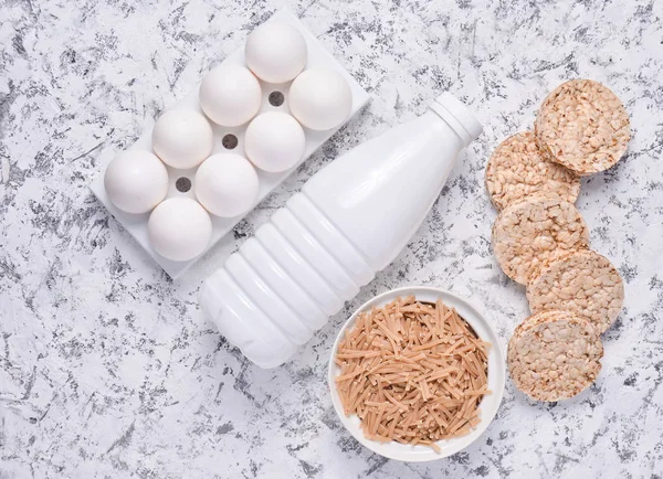 Top view of diet, healthy food. Crispy round dietary fitness bread, a bottle of yogurt, buckwheat noodles, plastic egg tray on a white concrete background. Flat la