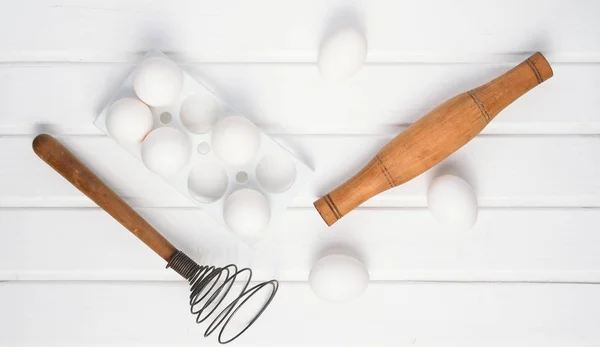 The cooking process. Plastic tray with white eggs, rolling pin, whisk on a white wooden table. Top view