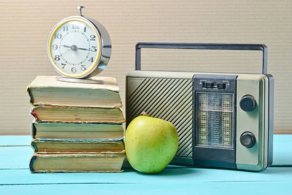 Alarm clock on stack of old books, radio receiver, apple on a blue wooden table. Retro still lif