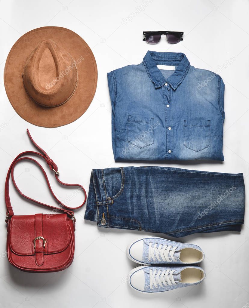 Women's fashion clothing and accessories. Jeans, denim shirt, sneakers, felt hat, leather bag, sunglasses, layout on a white background. Top view, flat la