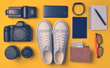 Becerikli ve aksesuarları düzen sarı bir arka plan üzerinde. Spor ayakkabı, fotoğrafik donanımlar, çanta ile dolar, akıllı saat, smartphone, defter, güneş gözlüğü. Seyahat, nesneleri, üst kavramı vie