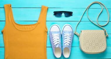 Kırmızı tişört, spor ayakkabı, deri çanta, mavi ahşap bir masa üzerinde güneş gözlüğü. Kadın giyim, Ayakkabı ve aksesuarları, üstten görünüm, la düz