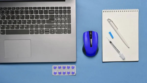 Workplace of a modern doctor. Laptop, wireless mouse, notebook, pills, syringe on a blue pastel background. Top view, minimalist tren