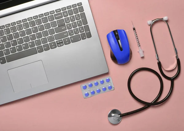 Workplace of a modern doctor. Laptop, wireless mouse, stethoscope, pills, syringe on a blue pastel background. Top view, minimalist tren