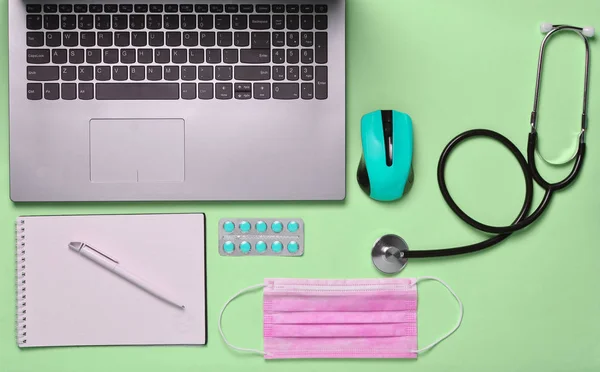 Workplace of a modern doctor. Laptop, wireless mouse, notebook, stethoscope, pills on a pink pastel background, top view, minimalist tren